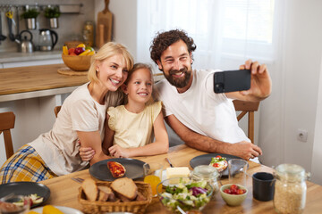 A family of three shares a meal at a dining table in a cozy kitchen. They smile for a selfie,...