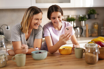A mother and daughter sit together at a wooden table in their home kitchen, laughing and looking at...