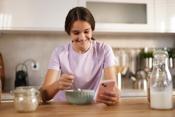 A teenage girl smiles while eating breakfast, holding a spoon in one hand and a smartphone in the...
