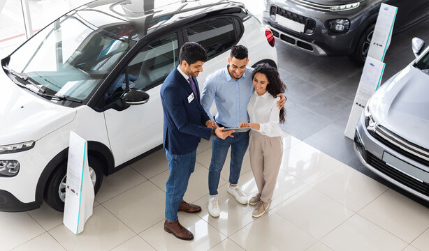 Top view of cheerful middle-eastern young couple having conversation with good-looking sales assistant at auto showroom, checking papers or car specification, full length photo
