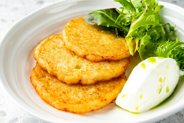 Golden brown potato pancakes served with sour cream and green salad on a white plate, top view,...