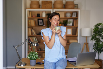 A woman sits on a desk in a cozy home office, smiling as she holds a coffee cup. The space is...