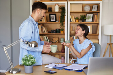 Inside a warm and inviting home office, two colleagues share a lighthearted moment over coffee,...