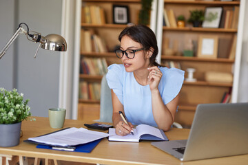 A person is actively writing in a notebook while seated at a desk in a tidy home office. Books are...