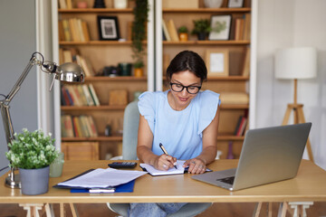 A woman sits at a tidy desk in a bright home office, writing notes in a notebook while surrounded...