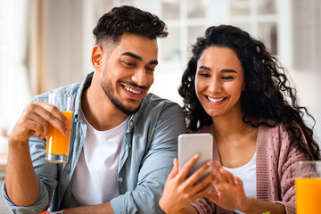 Portrait Of Cheerful Arab Lovers Using Smartphone During Breakfast In Kitchen, Young Happy Middle...