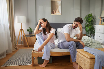 A couple sits on opposite sides of their bed, visibly distressed. The atmosphere suggests...
