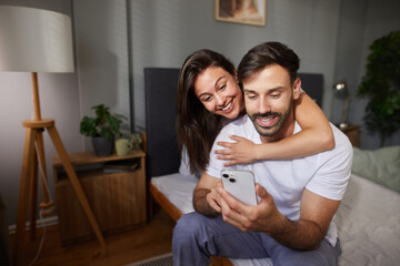 A couple sits on a bed, illuminated by soft light, sharing smiles as they look at a smartphone...