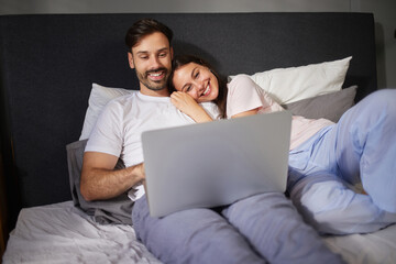 Two people relax in bed, smiling and engaging with a laptop. Soft lighting creates a warm...