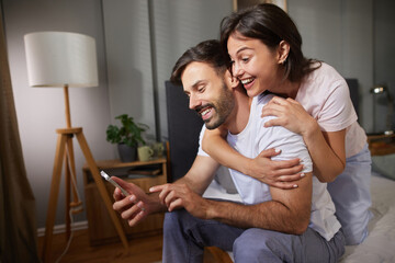 A man and woman sit together in their bedroom, engaged in a lighthearted moment as they share a...