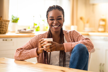 Cheerful young african american woman relaxing in cozy kitchen, drinking coffee. Joyful millennial...