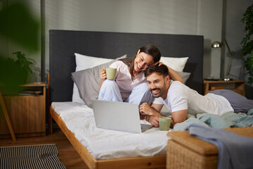 A couple sits together on their bed, smiling and browsing vacation options on a laptop. They hold...