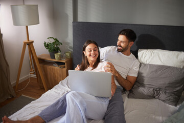 A couple is snuggled in bed, engaged with a laptop. They share laughter and a warm connection,...