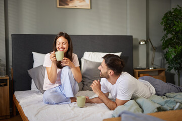 A couple sits comfortably on their bed, savoring steaming cups of coffee. They smile and engage in...