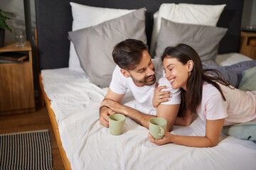 Two people are sitting on a bed, sharing a moment of joy while sipping coffee from casual mugs....