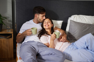 A couple sits together in bed, enjoying warm cups of coffee. They share laughter and affection in a...