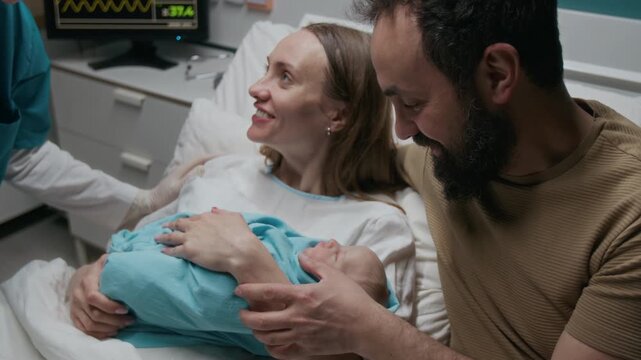 Medium close-up of joyous Caucasian couple, mother and father cradling newborn baby swaddled in blue sheet in delivery room in maternity ward after giving birth, admiring while talking to midwife