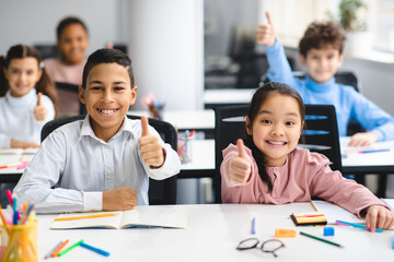 Education Concept. Diverse group of happy smiling international classmates sitting at desk in classroom and showing thumbs up sign gesture, girl and boy enjoying studying at school