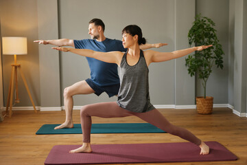 Mid adult man and woman practicing yoga together at home, doing the warrior ii pose