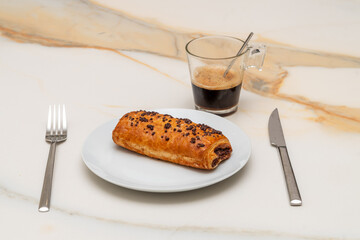 a white plate with a chocolate croissant on a white veined marble background with a stainless steel...