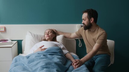 Medium full shot of loving middle-aged Caucasian man sitting on chair in hospital ward, stroking wife on hair, who is lying in bed, exhausted from labor contractions, sleeping and resting