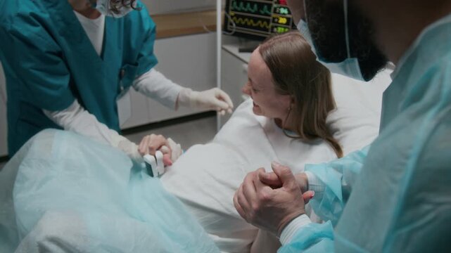 Medium shot of pregnant young Caucasian woman lying on bed in delivery room at maternity hospital, experiencing labor pain, screaming, breathing, while husband in holding hand and midwife counting