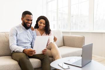 Portrait of cheerful african american couple sitting on the couch, man holding paper, working and...