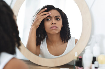 Fine lines and wrinkles. Concerned black woman looking in mirror checking aging signs on her face,...