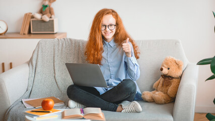 Portrait of positive smiling red-haired teenage girl in eyeglasses showing thumbs up gesture sitting on the couch and using laptop, doing homework, recommending online school or educational course