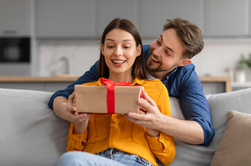 Excited young woman receiving surprise gift from man standing from back lady sitting on sofa,both...