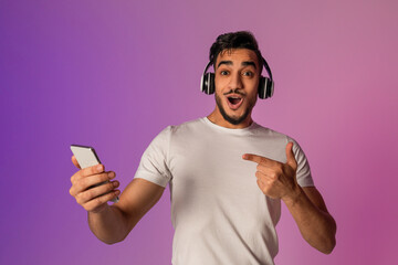 Surprised young Arab guy in headphones pointing at smartphone, shocked over online sale in neon...