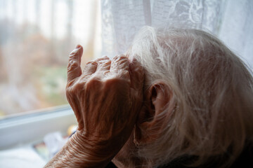 Grandma is sitting alone and looking out the window, in the frame her hand is near her head.