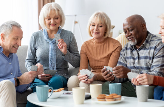 Multiracial group of happy senior people sitting on couch, playing cards, drinking tea, eating cookies at nursing home, enjoying time together. Residential facilities, assisted living concept
