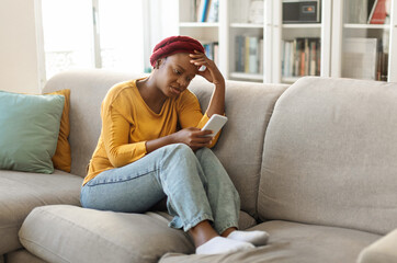 Obraz premium Breakup depression concept. Upset young black woman sitting on sofa with cellphone and reading bad message at home, female with mobile phone feeling sad, suffering from loneliness on web, copy space