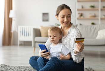 Millennial mother with baby boy using cellphone and credit card for online shopping at home, happy...
