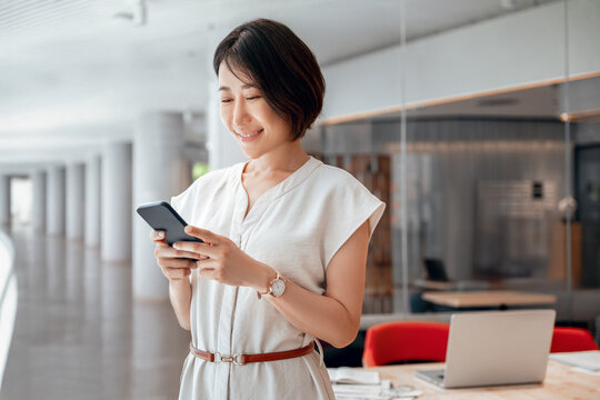 Asian mature business woman manager ceo using cell phone mobile app. Smiling eastern 40s years old female businesswoman holding smartphone in office workspace for trading, banking, working online
