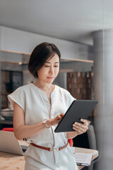 Naklejka na ściany i meble Middle-aged eastern businesswoman holding digital tablet using tab application standing at workplace in office. Mature Asian professional CEO business woman working on touchscreen computer. Vertical