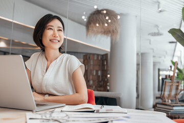 Middle-aged woman using computer technology for financial marketing work online. Dreaming mature Asian professional businesswoman analyst working on laptop pc in office, looking aside. Copy space