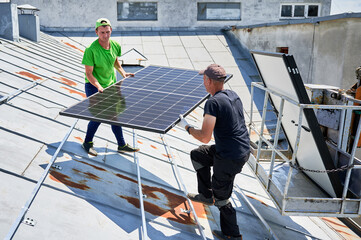 Workers building solar panel system on metal rooftop of house with assistance of crane lift. Two men installers carrying photovoltaic solar module outdoors. Renewable energy generation concept.