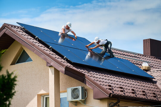 Workers building photovoltaic solar panel system on rooftop of house. Men technicians in helmets installing solar module with help of hex key outdoors. Alternative energy generation concept.