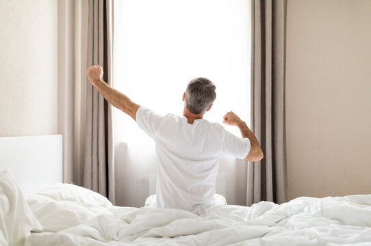 Rear view of unrecognizable middle aged man in pajamas sitting on bed and stretching body after waking up in the morning, looking at window, copy space. Comfortable healthy sleep concept - Powered by Adobe