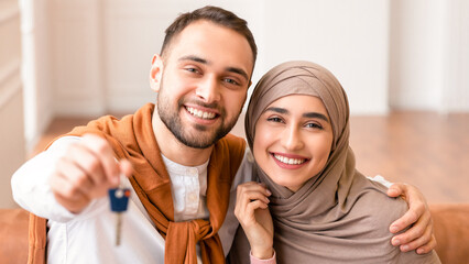 Own House. Cheerful Muslim Young Family Showing New Home Key Smiling To Camera Sitting On Sofa In...