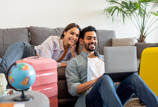 Happy loving couple planning trip after quarantine, buying low price tickets and booking hotel online, using laptop while sitting in living room with packed suitcases nearby