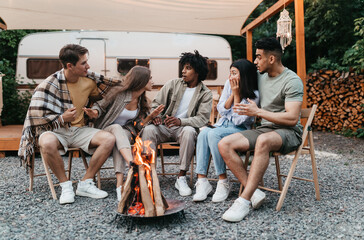 Diverse young friends sitting near campfire, telling scary stories next to motorhome during camping...