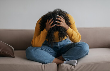 Depressed young black woman sitting cross legged on sofa with head in her hands at home. African...