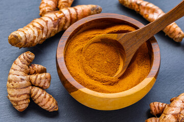 Turmeric roots and powder in a wooden bowl on a dark slate board: natural superfood and culinary spice