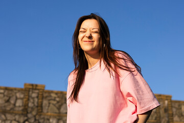 Teen girl in pink t-shirt smiling, squinting under sun on blue sky background.