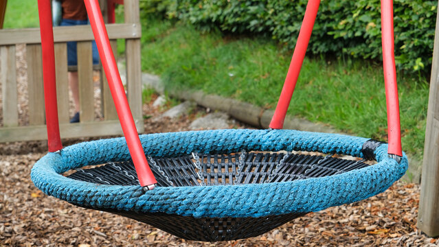 A modern round rope swing with a blue edge and red metal supports sits in a playground, surrounded by wood chips and green grass.
