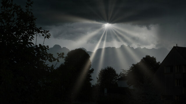 Dramatic sunbeams pierce through dark storm clouds, casting radiant light over silhouetted trees and rooftops against a mountainous backdrop.
