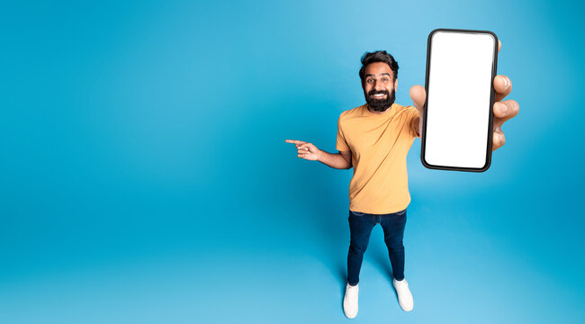 Mobile offer. Happy indian man showing phone with blank screen and pointing finger aside at free space on blue studio background, panorama, mockup. Choose this application